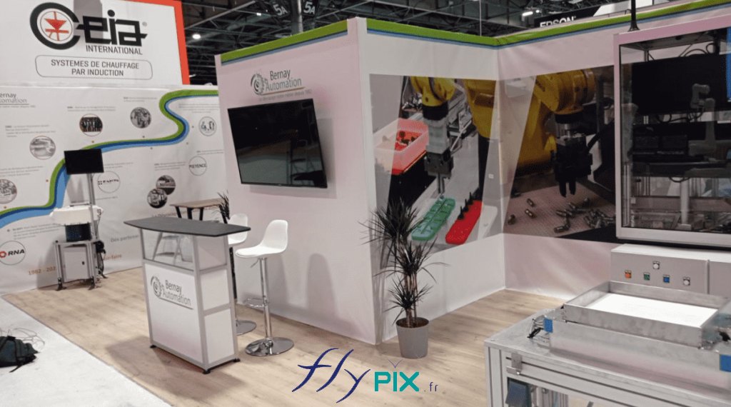 BERNAY AUTOMATION : vue générale présentant le stand modulaire, déployé pendant le salon professionnel, avec les différents composants comme le comptoir, le mobilier, les chaises, les plantes, la TV, les grands panneaux PLV imprimés, le sol en lino effet parquet bois.
