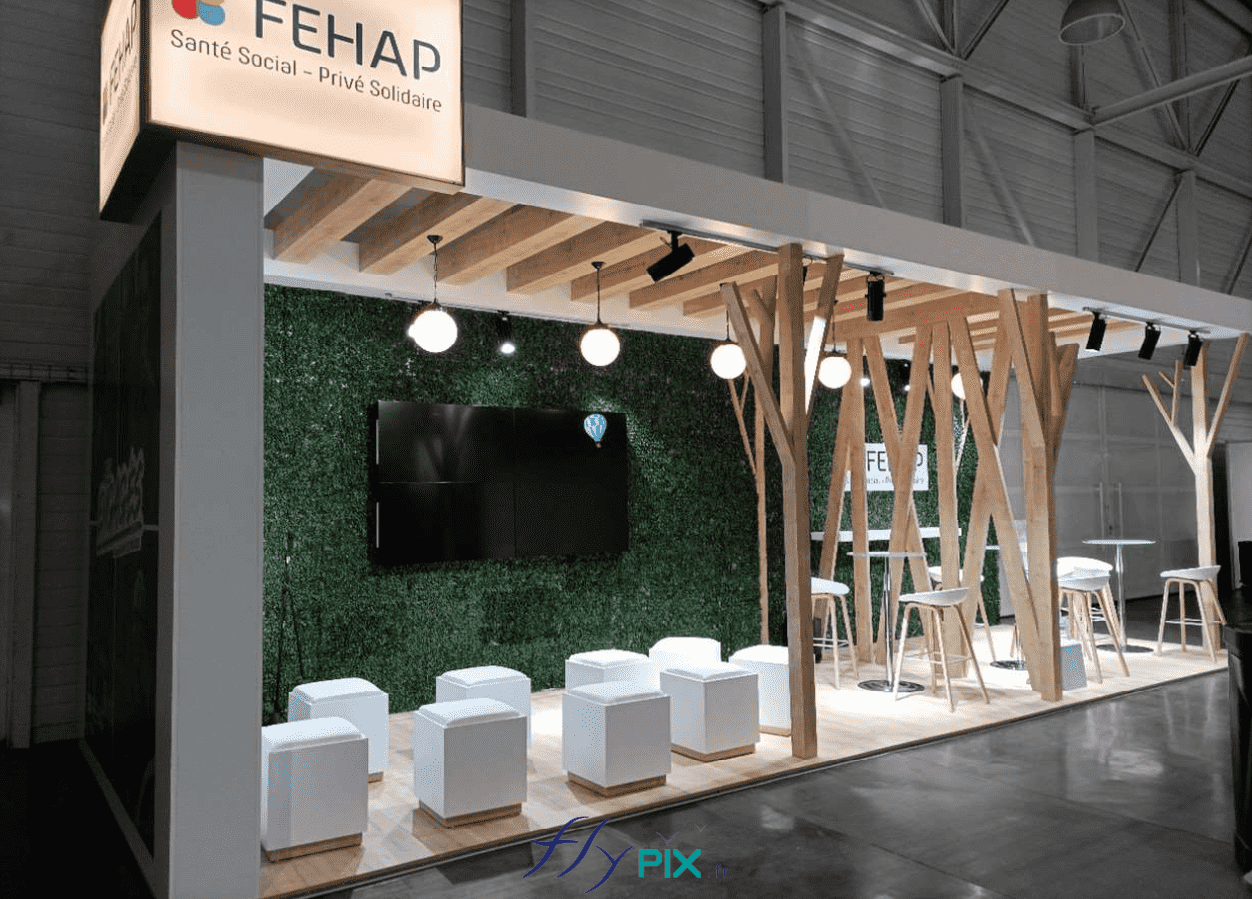 FEHAP : vue générale du stand de salon professionnel, en menuiserie : sol lino effet bois, tables, chaises, mobilier, lampadaires, lampes, structures en bois, fond en faux mur végétalisé.