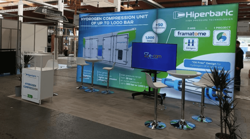 HIPERBARIC : vue générale d'un stand modulaire, une fois posé et installé par nos soins, avec en arrière-plan un grand panneau PLV imprimé (infographies), au sol un lino effet parquet bois, un comptoir visible au premier plan, du mobilier (tables et chaises) et aussi une télévision.