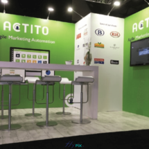 ACTITO : un bel exemple de stand de salon professionnel, de type modulaire, avec TV, télévision, sol lino parquety bois, comptoir, table, chaises, mur grands panneaux PLV imprimés et logotés, sur fond vert.