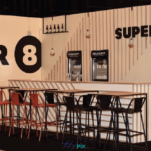 SUPER 8 : un stand de salon professionnel de type modulaire, avec un très grand comptoir à bar pour servir des boissons, des tables et des chaises, au fond des grands murs de panneaux PLV imprimés avec des gros logos sur fond blanc.